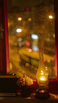 Warm indoor scene with an oil lamp on a windowsill, rain-dropped window and city lights outside.