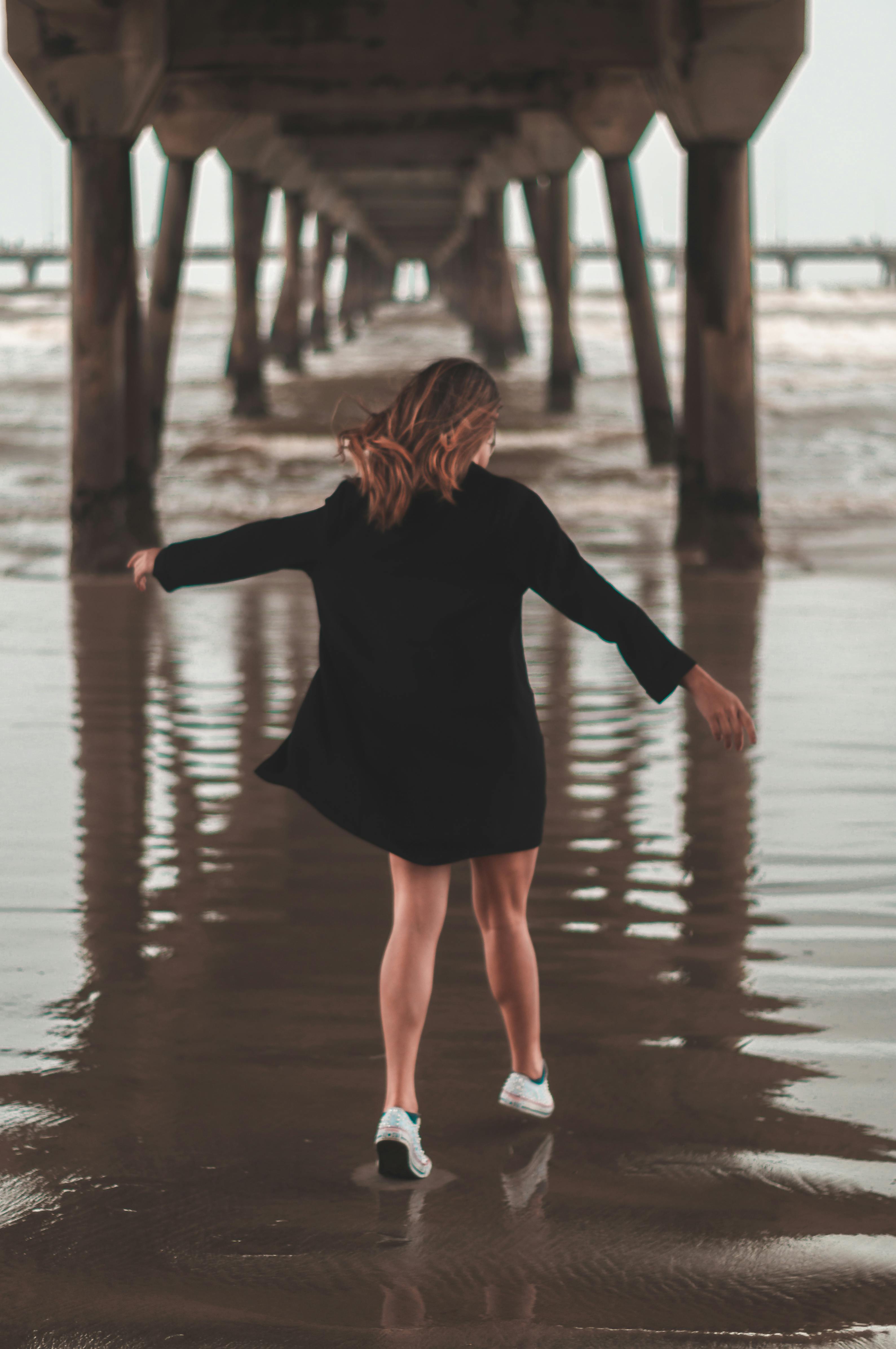 一名身穿黑色连衣裙的女子，在海滩的码头下无忧无虑地散步，尽显自由