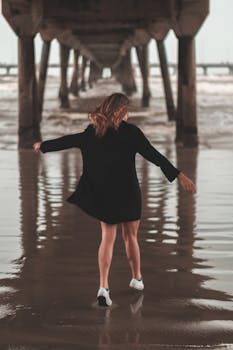 一名身穿黑色连衣裙的女子，在海滩的码头下无忧无虑地散步，尽显自由
