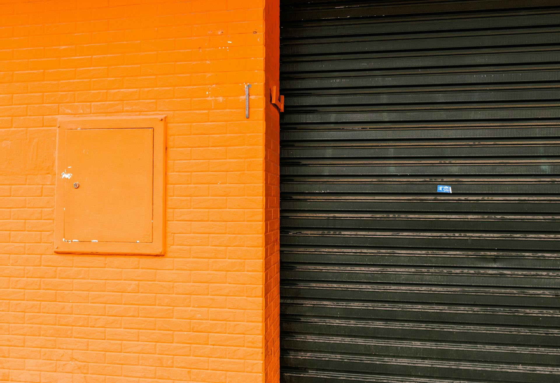 An orange-painted wall with imitation brick texture and a contrasting black roller door in an urban setting.