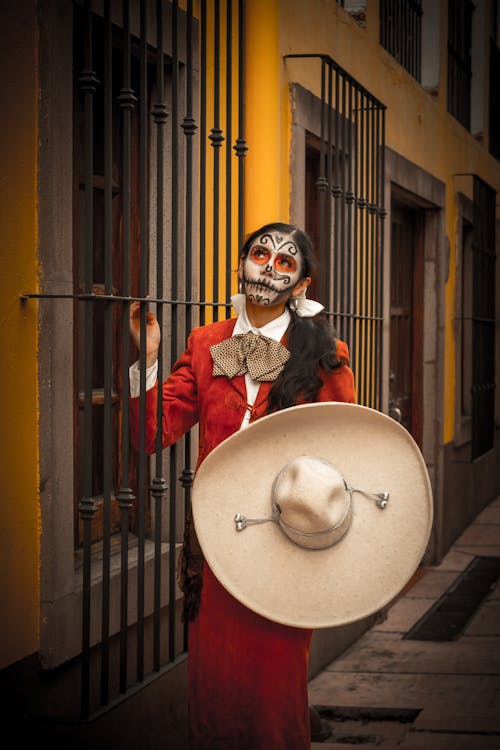 Woman with Sombrero and Face Painted as Catrina Standing by Bars on Window on Building Wall