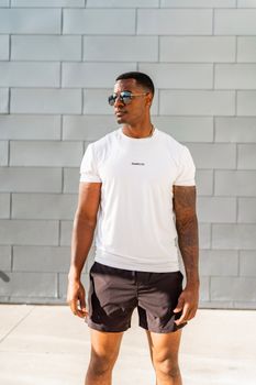 A man in a white shirt and sunglasses poses outdoors against a light brick wall.