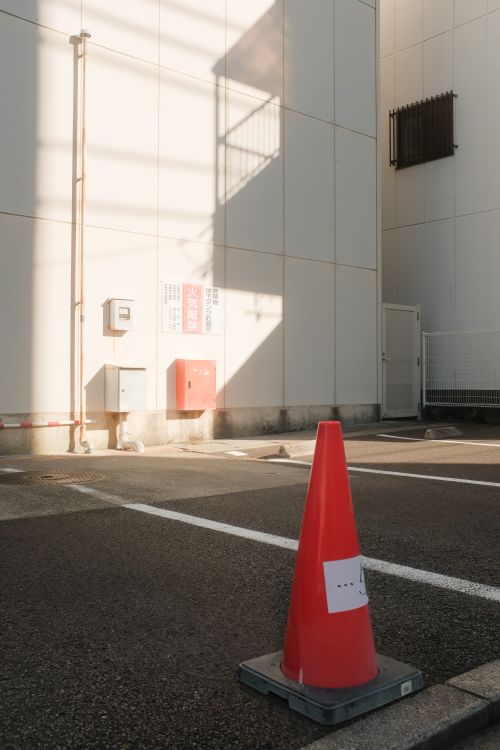 Free A red traffic cone sits in a sunlit urban parking lot with building shadows. Stock Photo