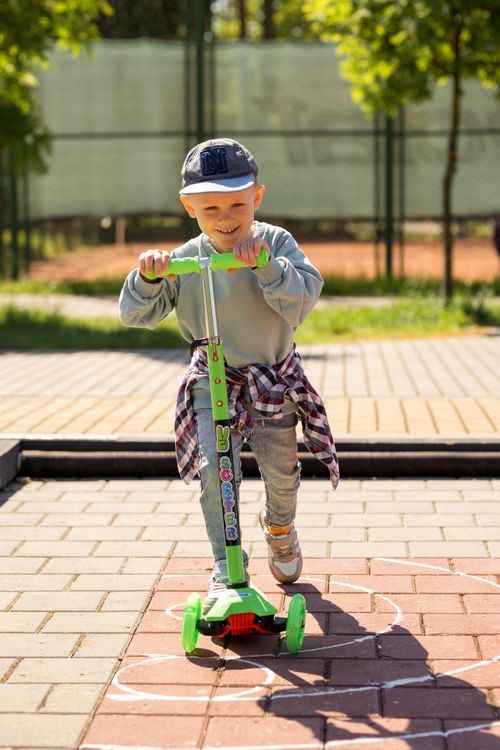 Free A happy child energetically rides a green scooter in a sunny outdoor setting, showcasing fun and playfulness. Stock Photo