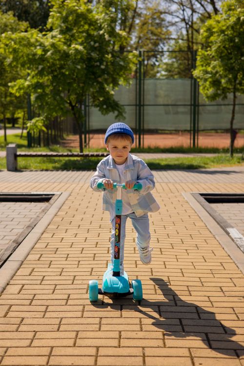 Free A young child joyfully rides a scooter on a sunlit path in an urban park. Stock Photo