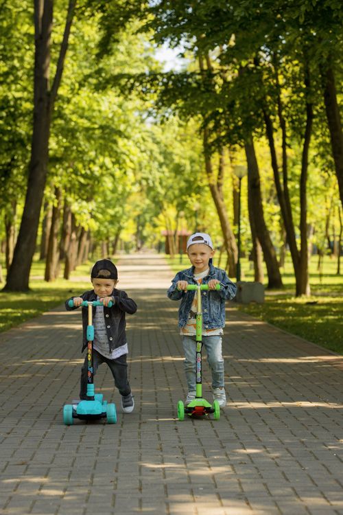Free Two young boys having fun riding scooters on a sunny park pathway, enjoying a day outdoors. Stock Photo