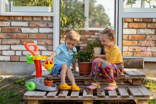 Free Two children sitting on wooden pallets, playing with toys, and watering plants. Bright and joyful outdoor scene. Stock Photo