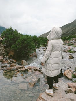 A woman in a warm jacket stands by a flowing stream amidst rocky, mountainous terrain in Cusco, Perú.
