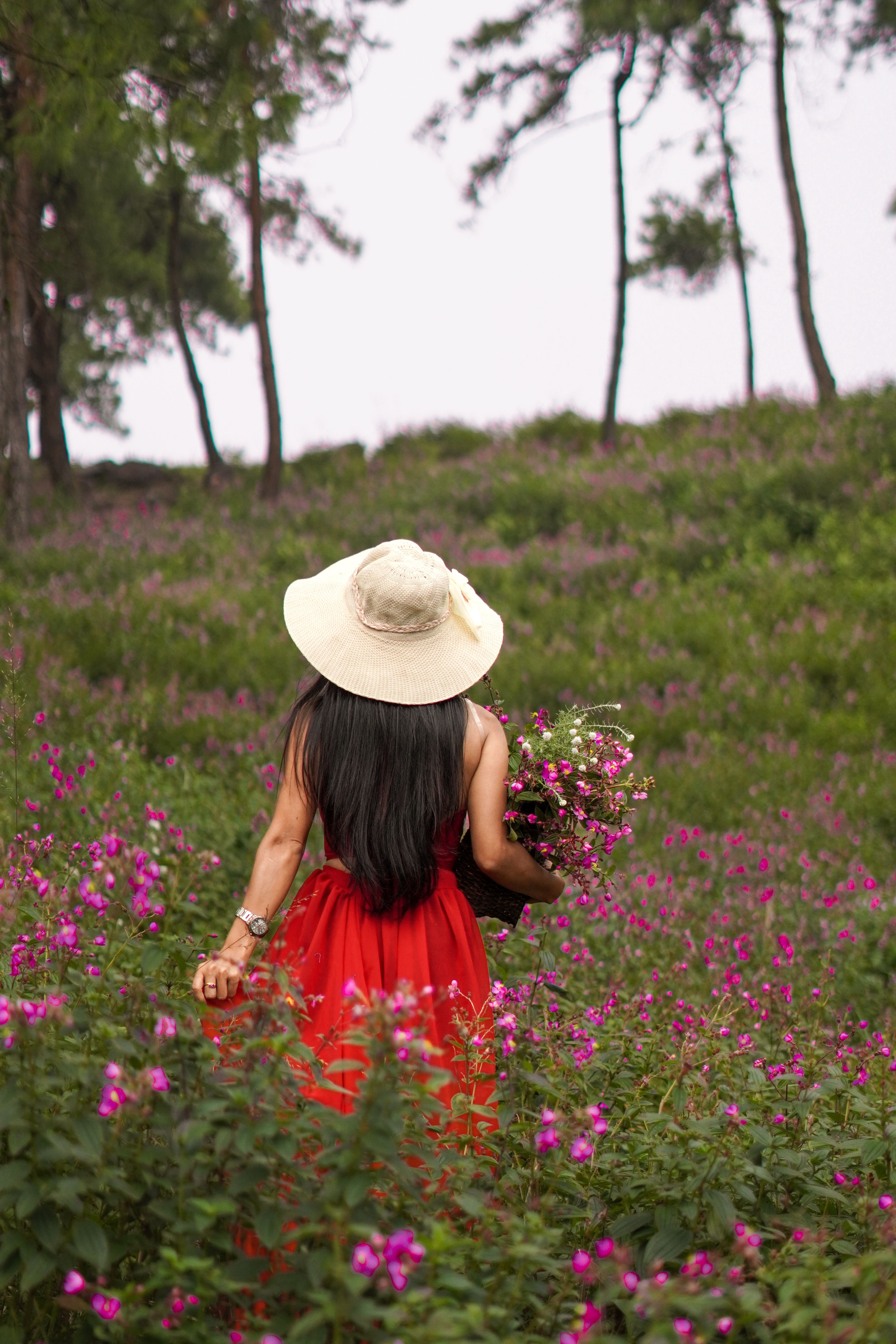 A woman in a red dress and hat holds flowers while walking through a vibrant meadow of purple blooms.