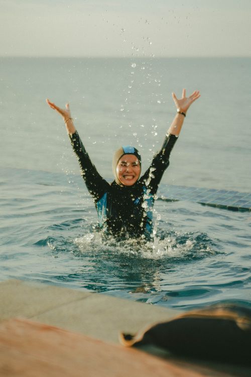Free Smiling woman in a hijab enjoys a swim, hands raised in celebration. Stock Photo
