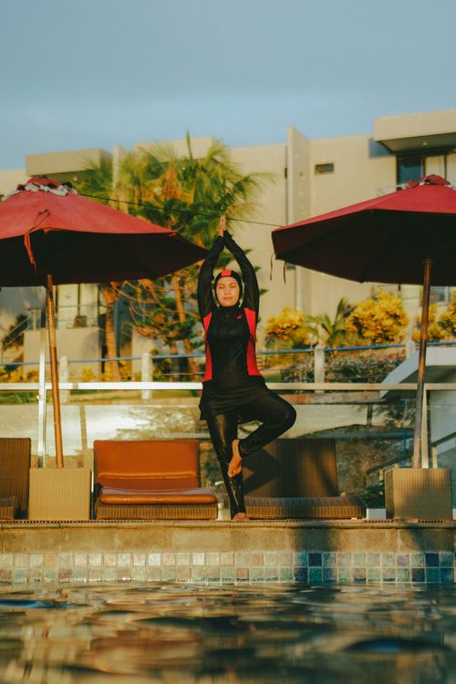 Free A woman in swimwear performs yoga by a pool, surrounded by umbrellas and deckchairs. Stock Photo