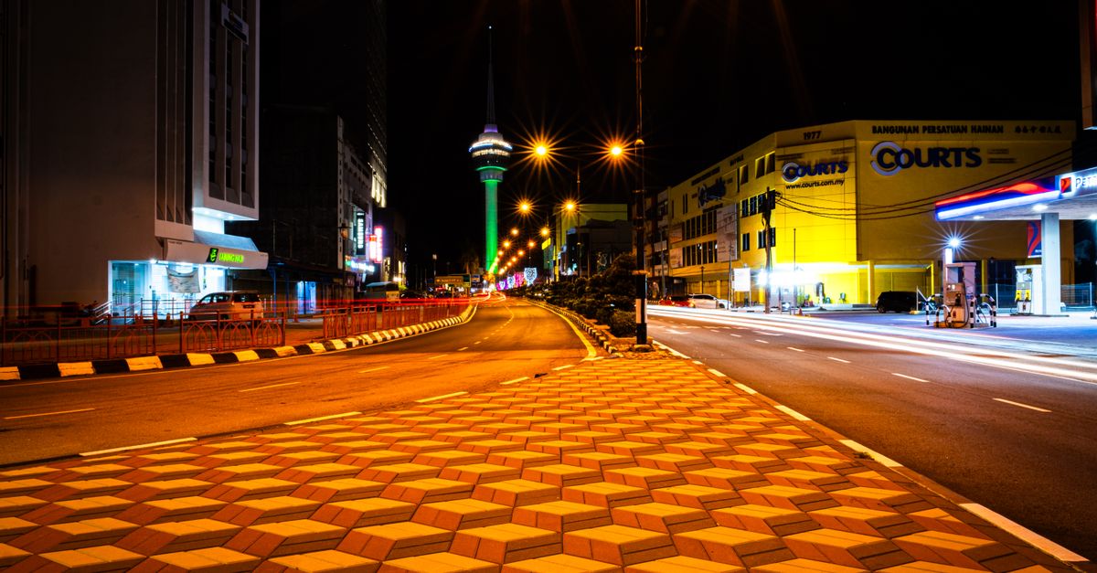 Nighttime city street with geometric sidewalk and illuminated tower in the background.