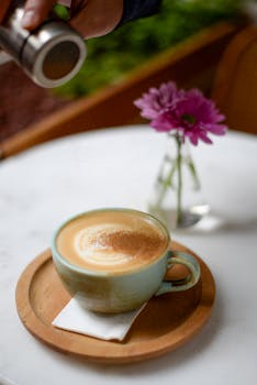 Close-up of a coffee latte with sprinkled cinnamon beside a vase of flowers, on a marble table.
