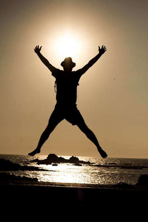Silhouette of Man Jumping with Arms Raised on Sea Shore at Sunset