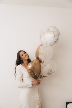Smiling bride in a white dress holding elegant gold and white wedding balloons indoors.