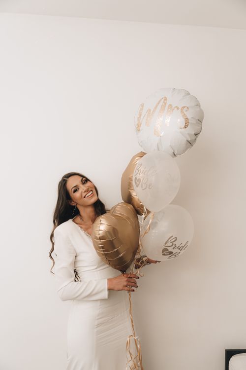 Free Smiling bride in a white dress holding elegant gold and white wedding balloons indoors. Stock Photo