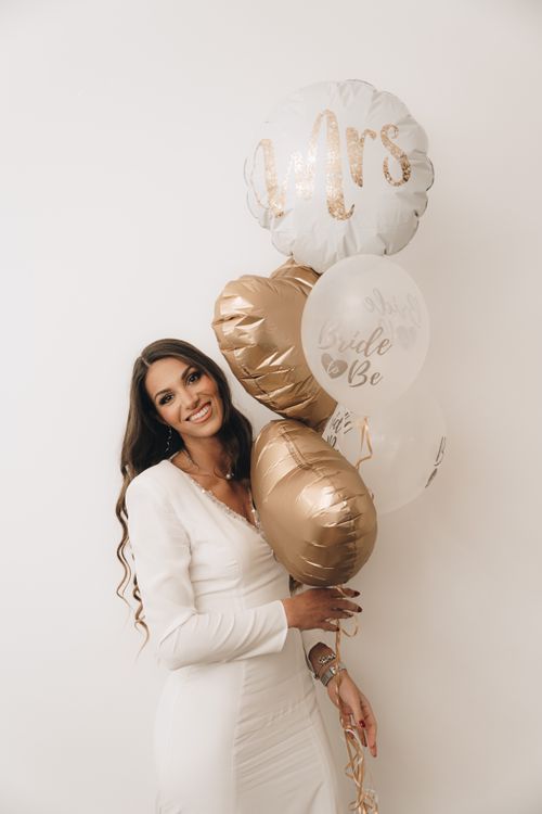 Free A smiling bride-to-be in a white dress holding elegant gold and white balloons indoors. Stock Photo