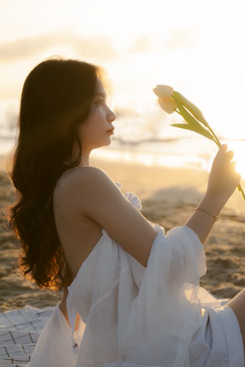 Woman in White Clothes Sitting with Flowers on Beach