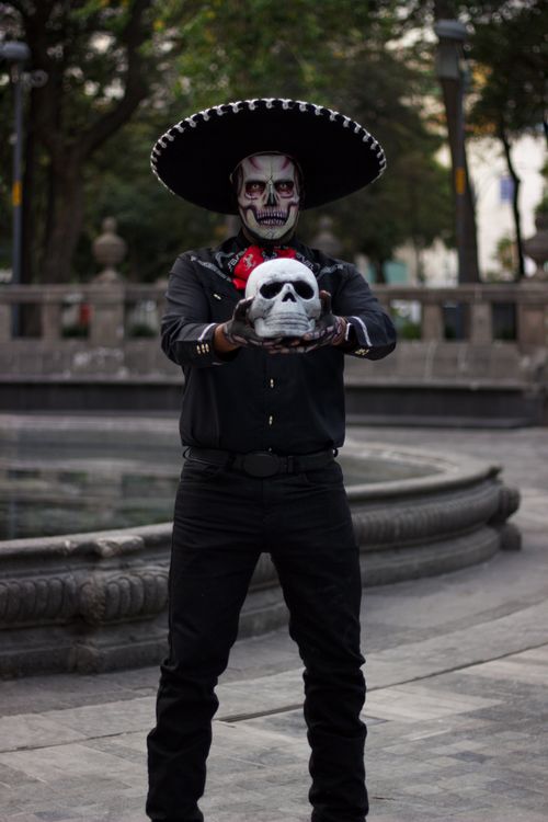 Free A man in traditional Mexican attire with a painted skull face holding a decorative skull outdoors. Stock Photo