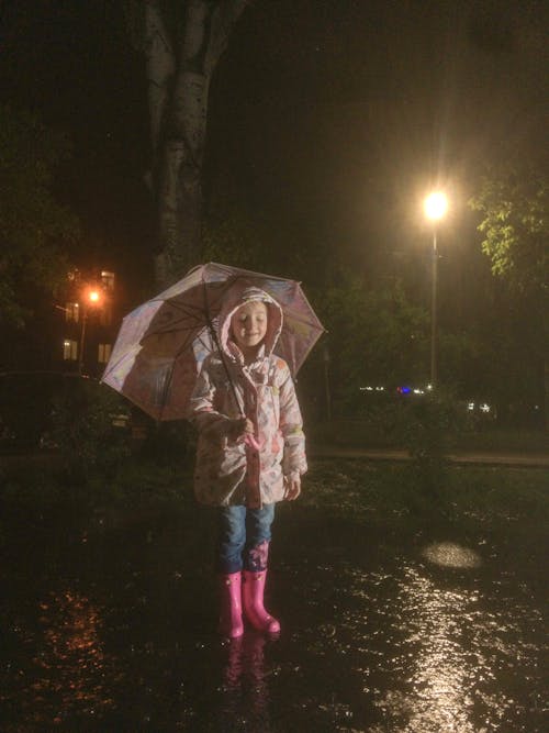 Delight in the image of a happy kid, adorned in stylish pink boots and a vibrant umbrella, relishing playful moments under the rain at night. Explore the joy of a child's rainy nighttime a...