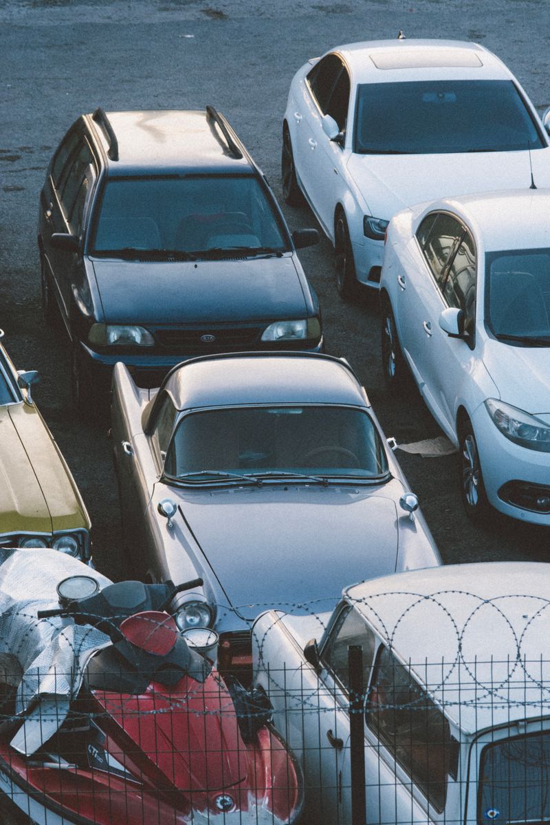 A mix of classic and modern cars parked in an outdoor lot behind a barbed wire fence.