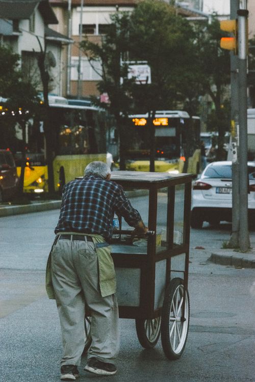 Free An elderly man pushes a street food cart on a bustling urban street, with traffic around him. Stock Photo