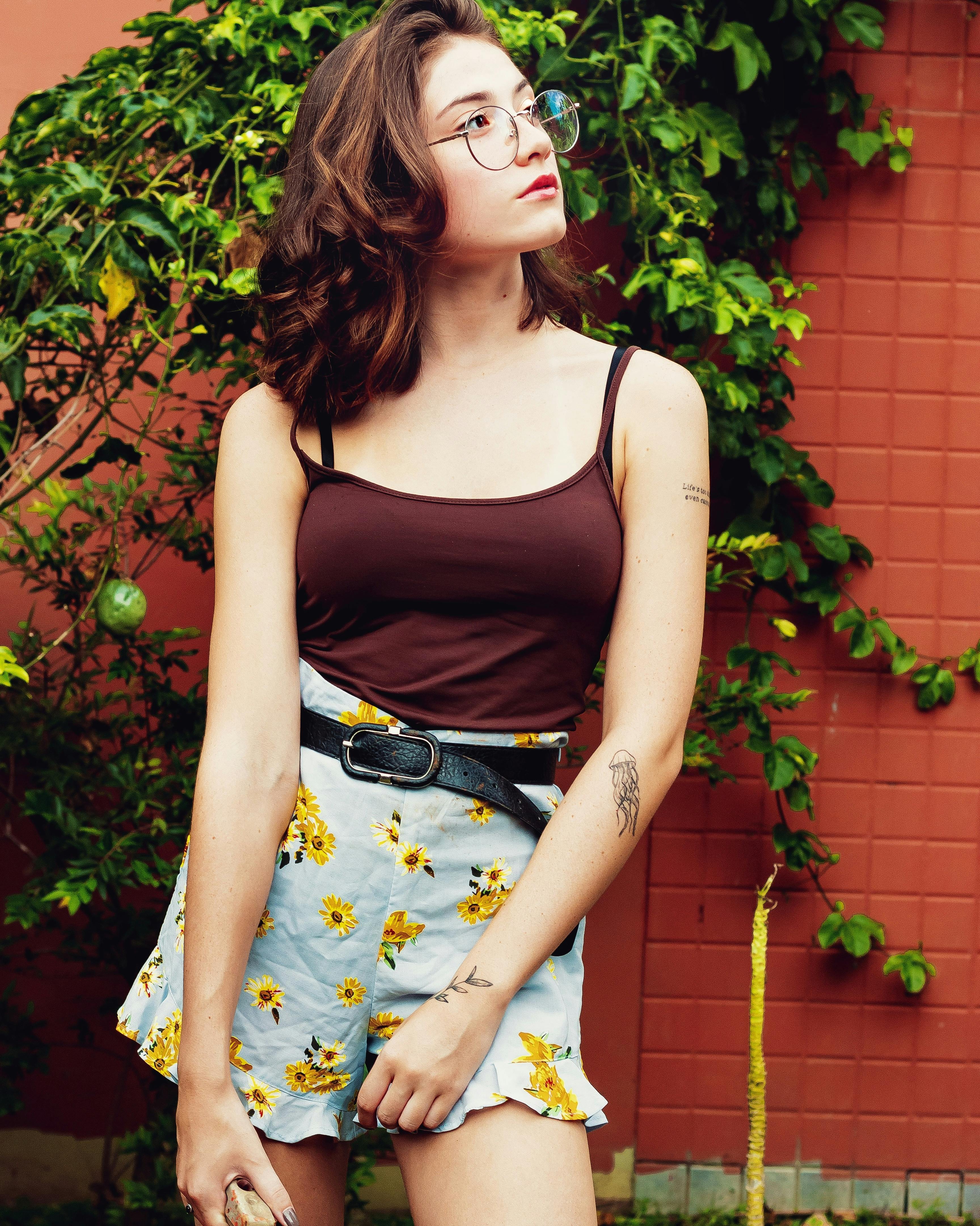 一位戴着眼镜、身穿花卉裙子的时尚年轻女子，在户外倚着红墙摆姿势