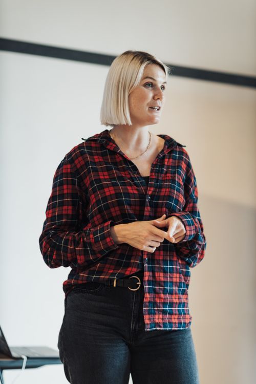 Free Blonde woman standing indoors, giving a presentation in casual attire, with a focused expression. Stock Photo