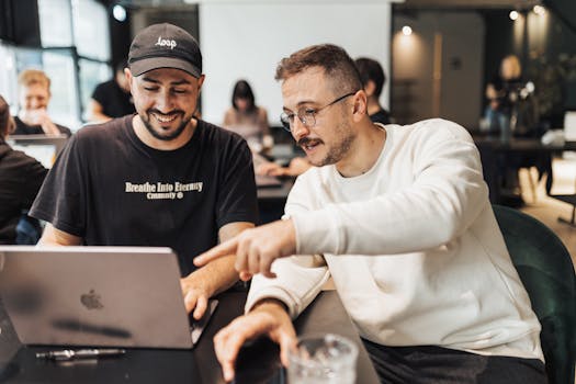 Two young men discussing project ideas while working on a laptop in a contemporary office setting.