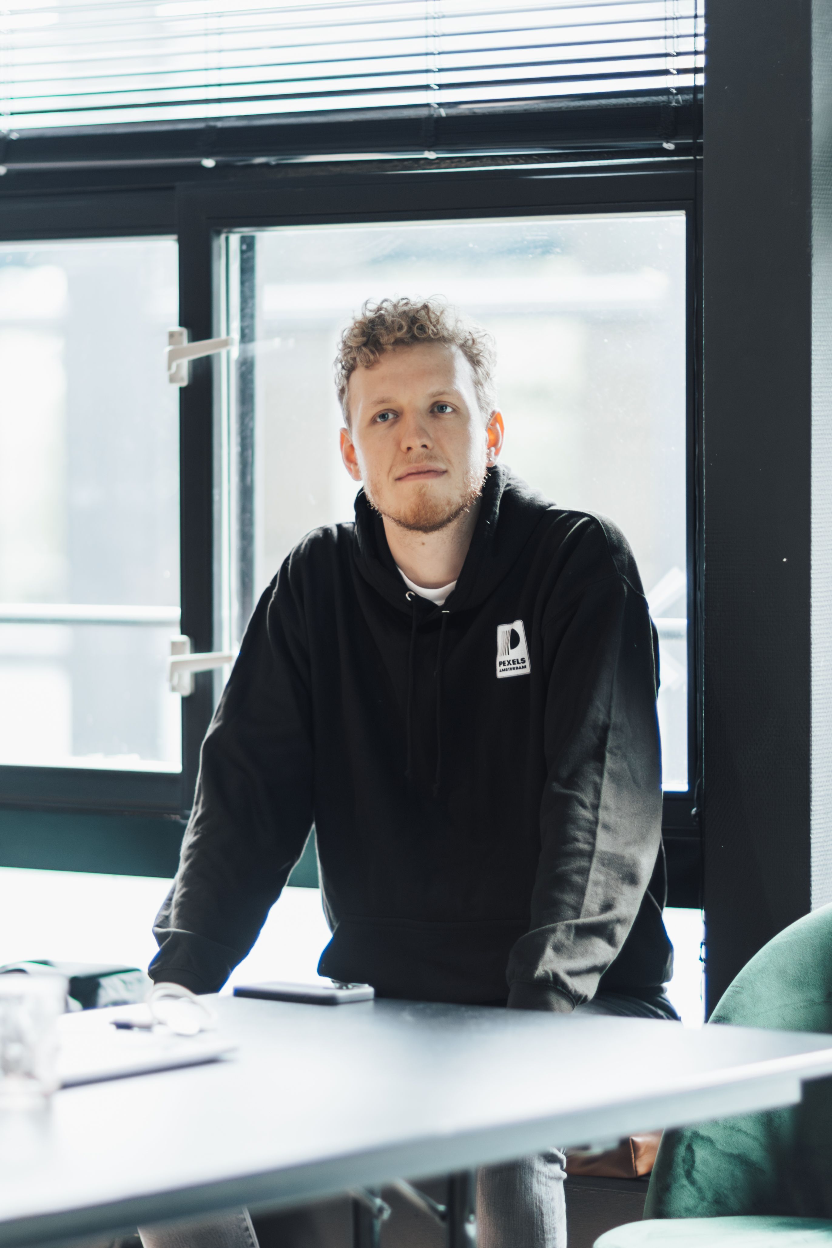 A man in a black hoodie is sitting by a window indoors, looking thoughtful and relaxed.