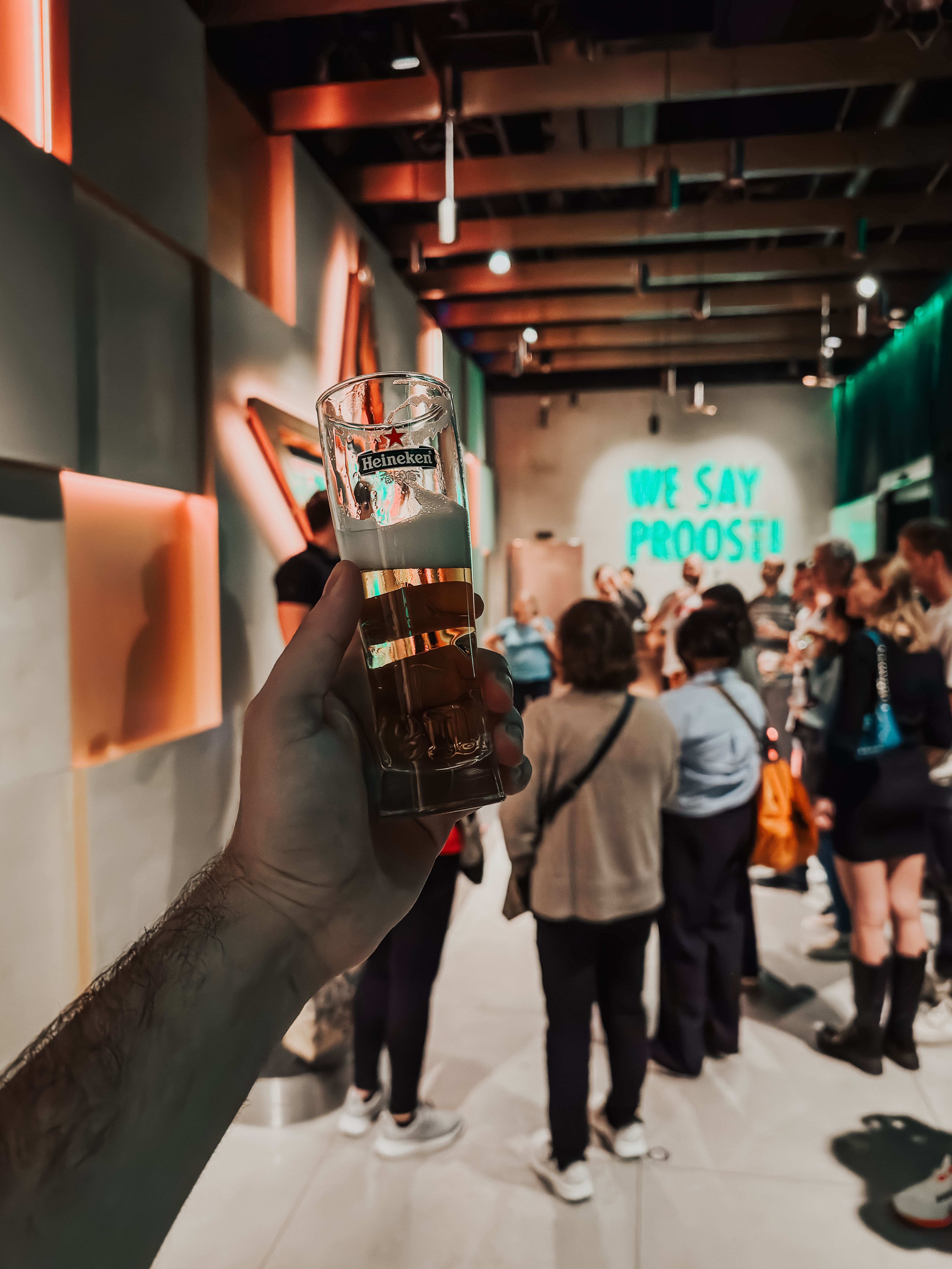 Close-up van een hand die een bierglas vasthoudt tijdens een brouwerijtour in Amsterdam met een levendige groep op de achtergrond.