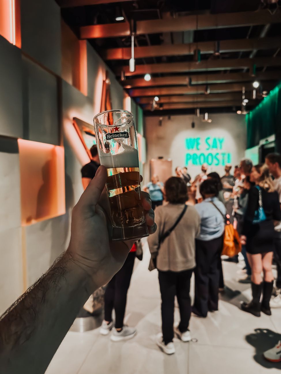 Close-up van een hand die een bierglas vasthoudt tijdens een brouwerijtour in Amsterdam met een levendige groep op de achtergrond.