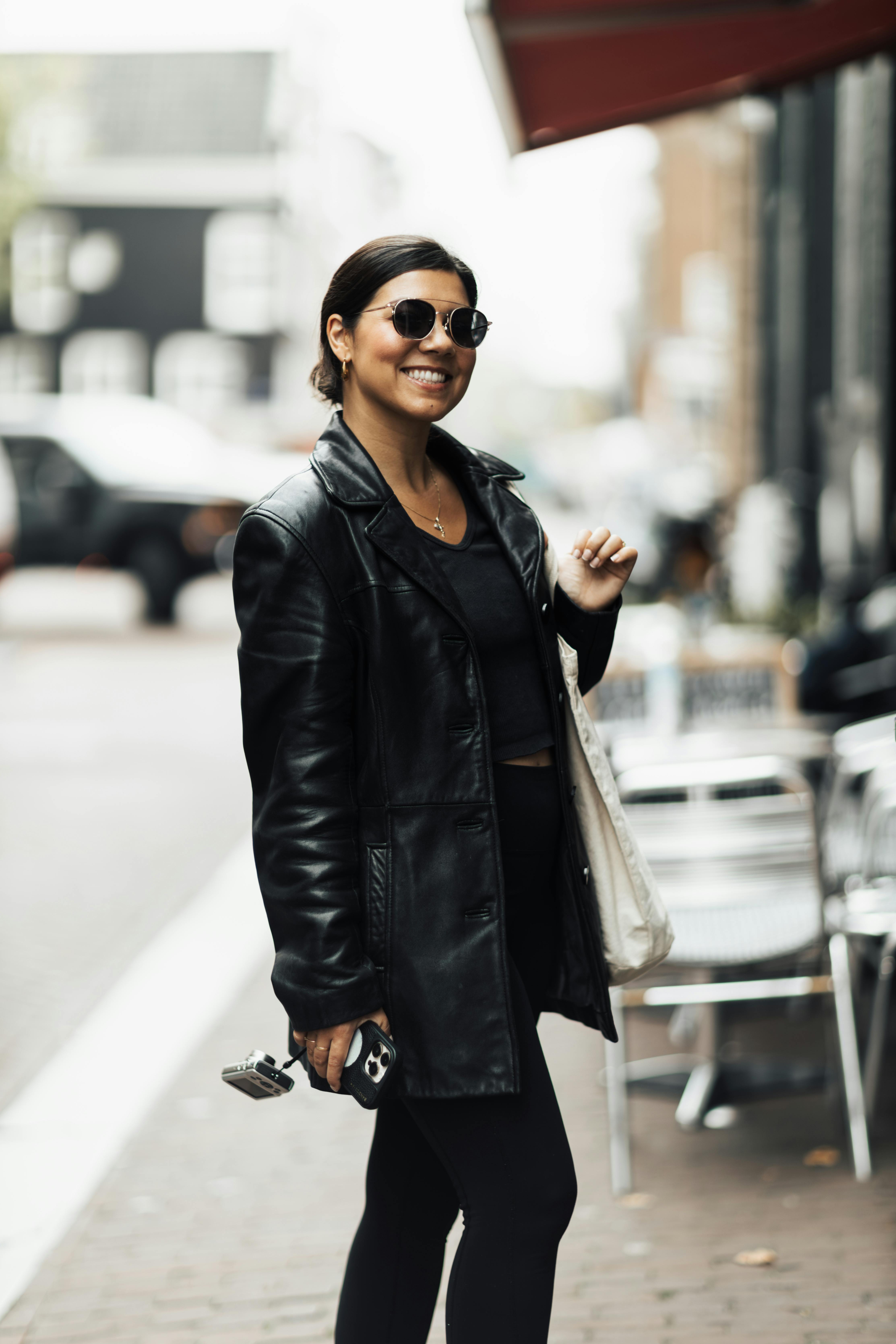 Smiling woman in a black leather coat and sunglasses standing on a bustling city street.