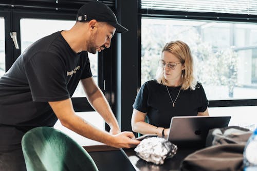 Blonde Woman and Man in Cap and T-shirt by Table with Laptop