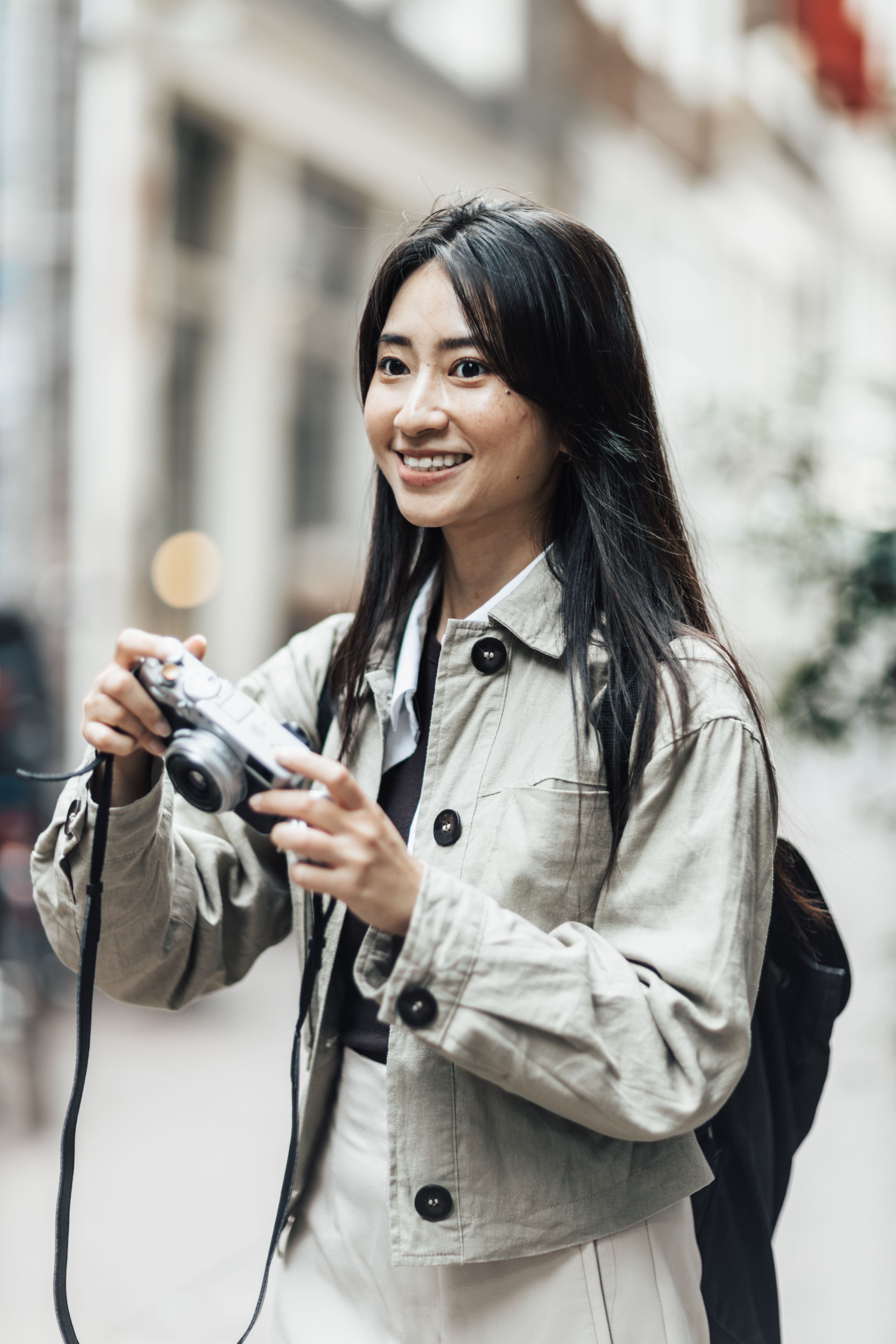 Asian woman holding a camera and smiling while exploring urban city streets, perfect for tourism visuals.