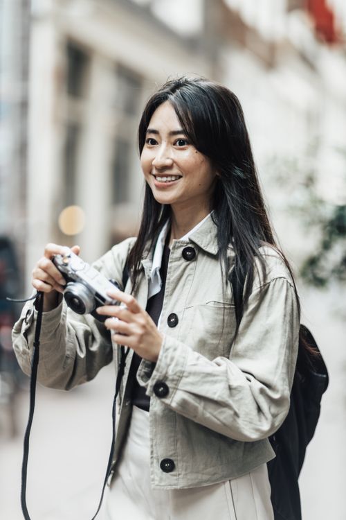Free Asian woman holding a camera and smiling while exploring urban city streets, perfect for tourism visuals. Stock Photo