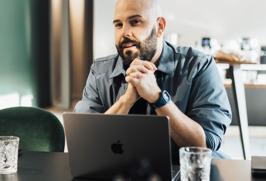 Um homem de barba, com as mãos entrelaçadas diante do notebook, escuta com atenção durante uma conversa de trabalho.