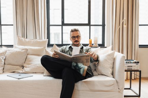 Free A man relaxes on a sofa, engrossed in a book, in a stylish, well-lit living room. Stock Photo