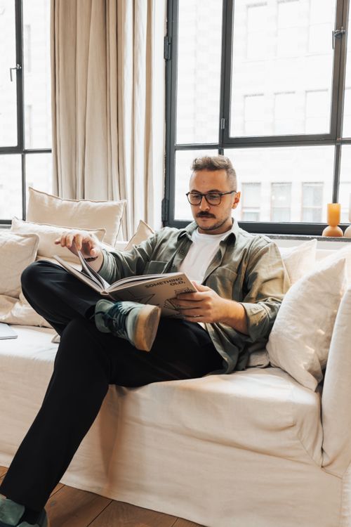 Free Man reading a book on a sofa by the window, enjoying a relaxed afternoon indoors. Stock Photo