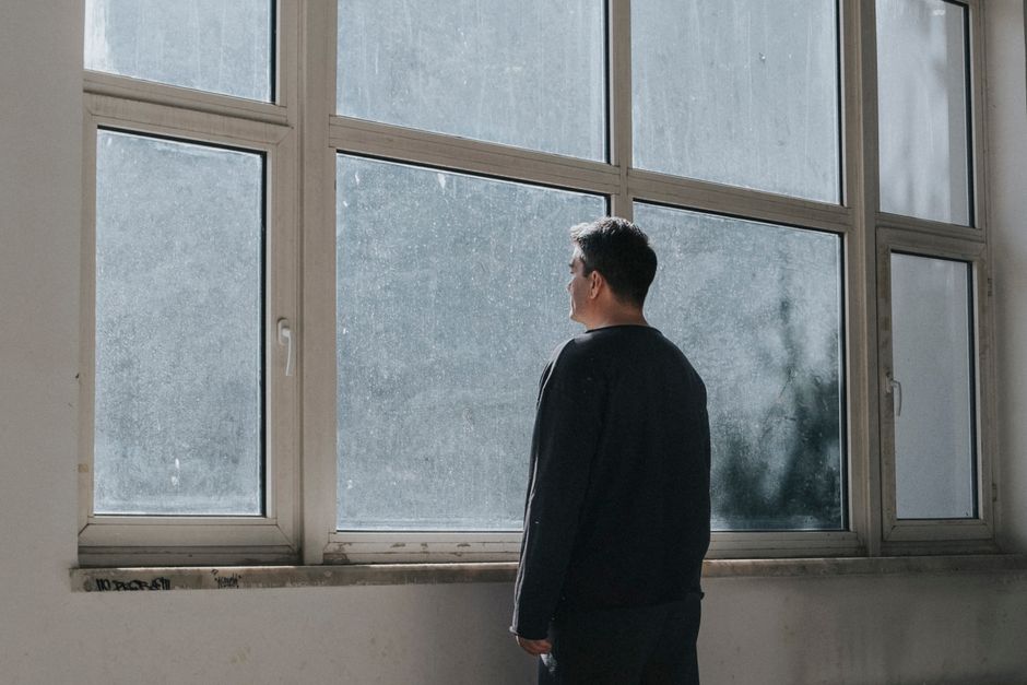 An adult man stands indoors staring contemplatively out dirty windows.