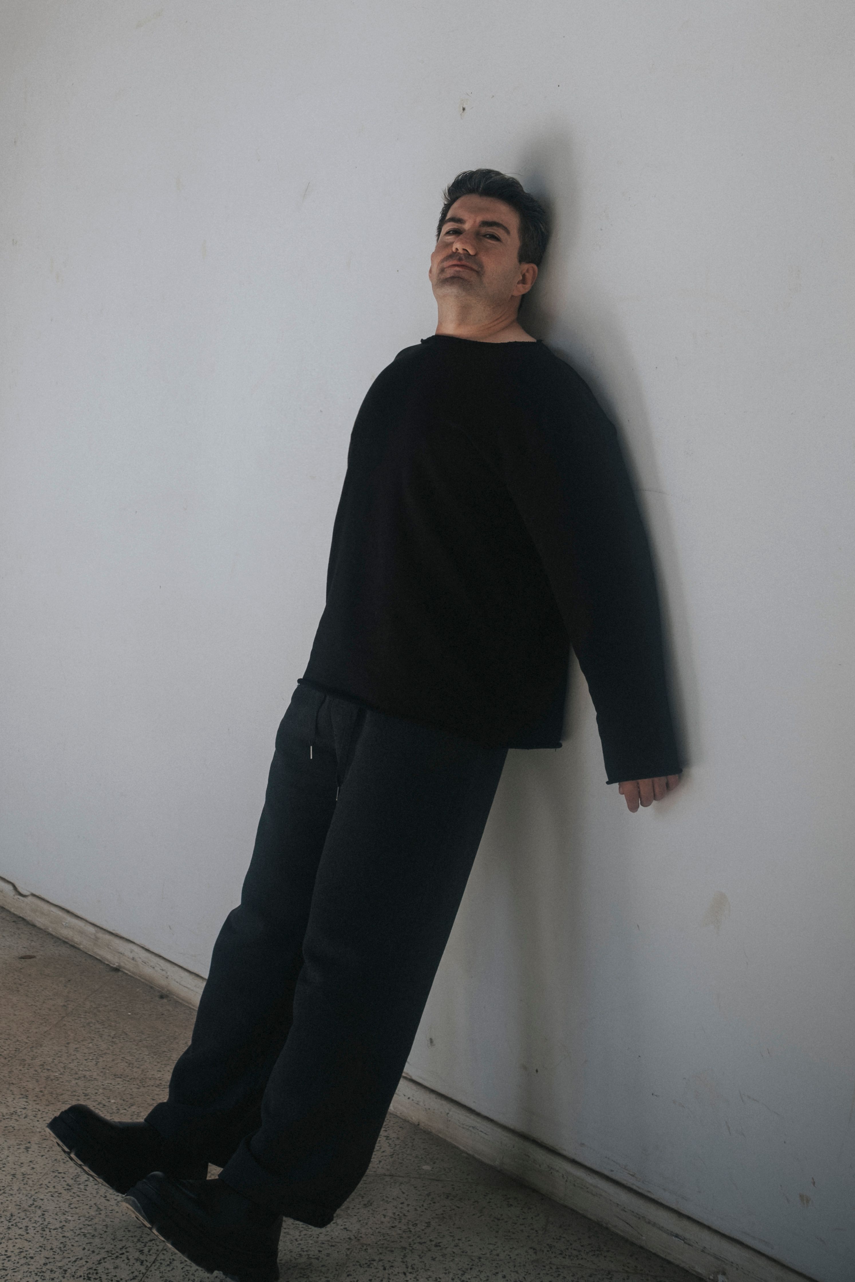 Adult man in casual attire leaning against a white wall indoors.