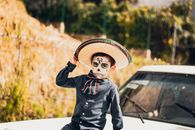 Child in traditional Day of the Dead costume with face paint and sombrero outdoors.