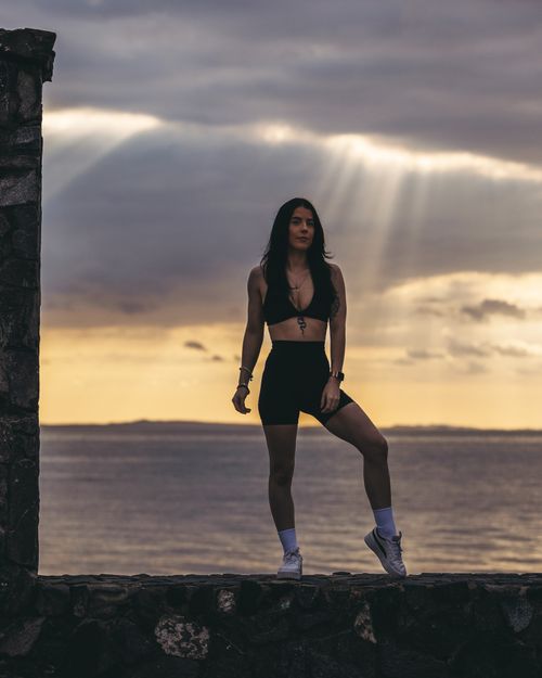 Free A woman in athletic wear poses confidently against a stunning ocean sunset backdrop. Stock Photo