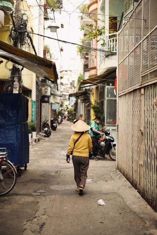 Free A person wearing a traditional conical hat walks through a bustling urban alleyway. Stock Photo