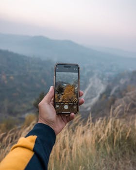 A hand holds a smartphone capturing the scenic autumn views of Srinagar's hills.