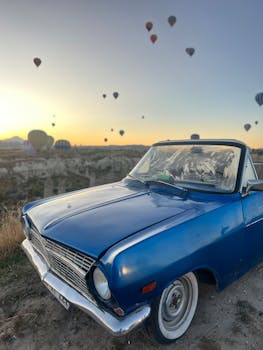 A classic blue convertible parked with hot air balloons over Cappadocia's landscape at sunrise.