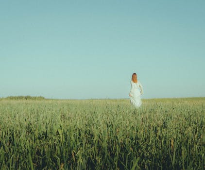 A woman in a white dress walks through a lush green field under a clear summer sky.