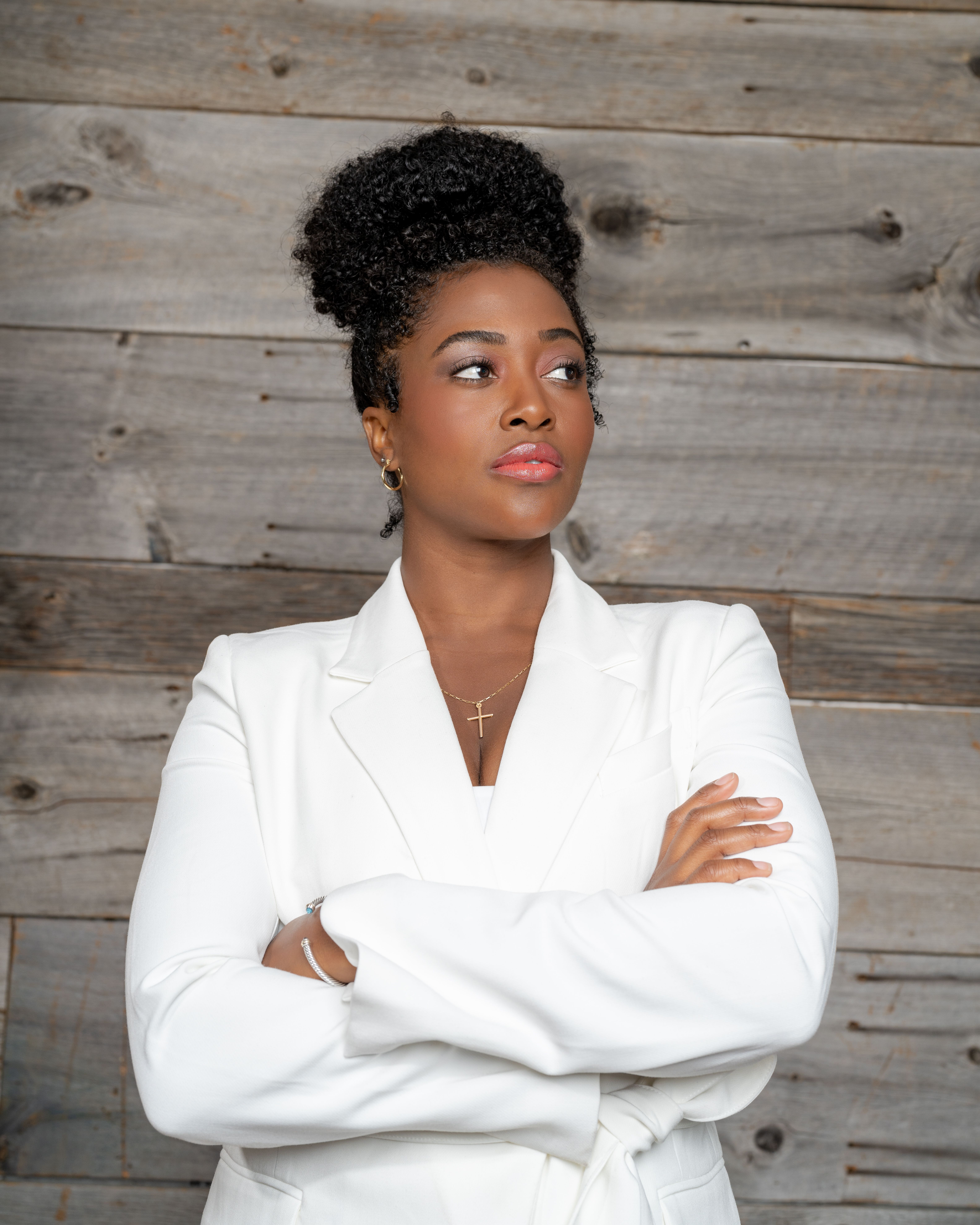 Businesswoman posing confidently with arms crossed wearing a stylish white suit against a rustic wooden background.