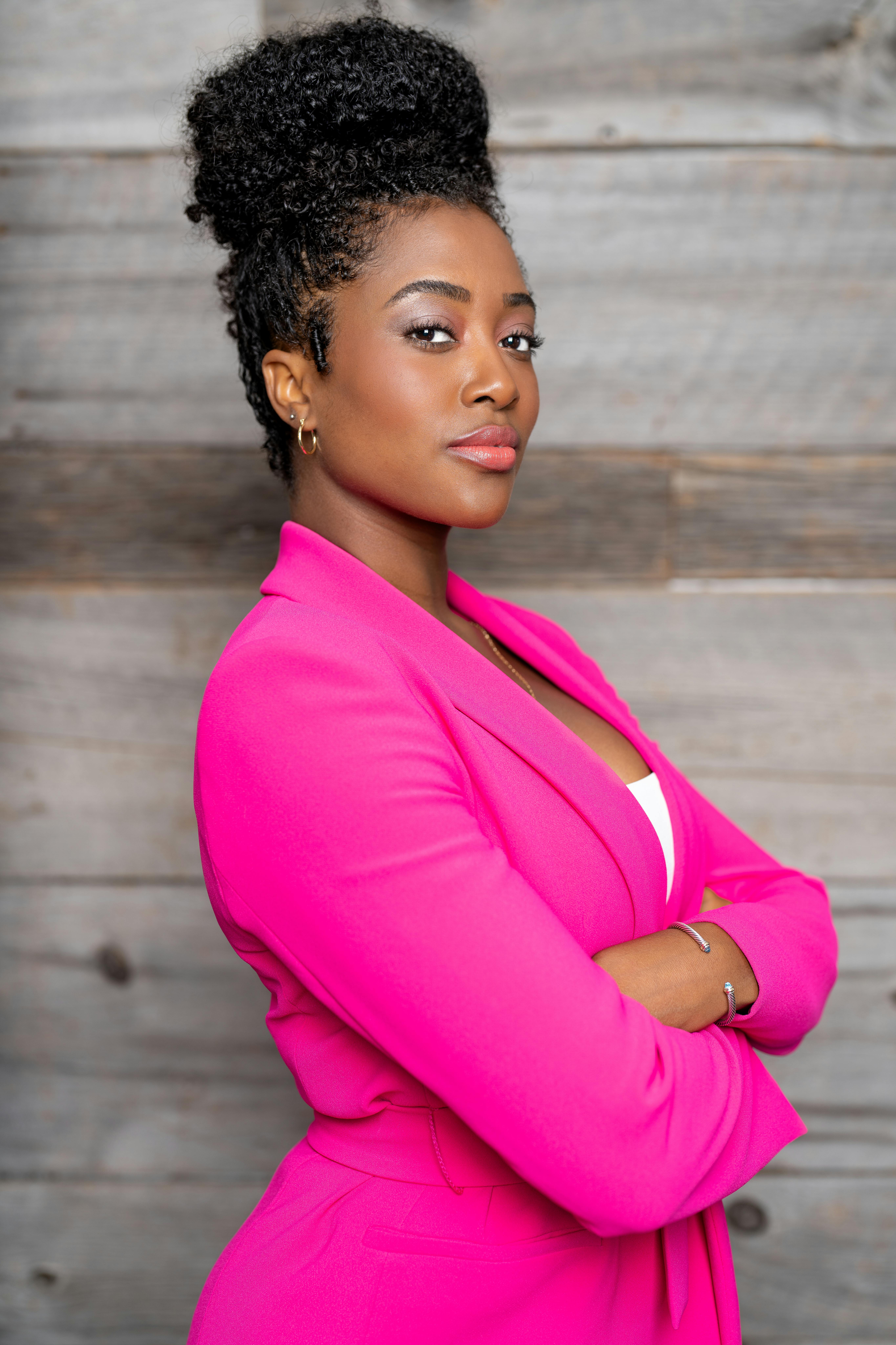 Black woman in a stylish pink jacket poses confidently against a wooden background.