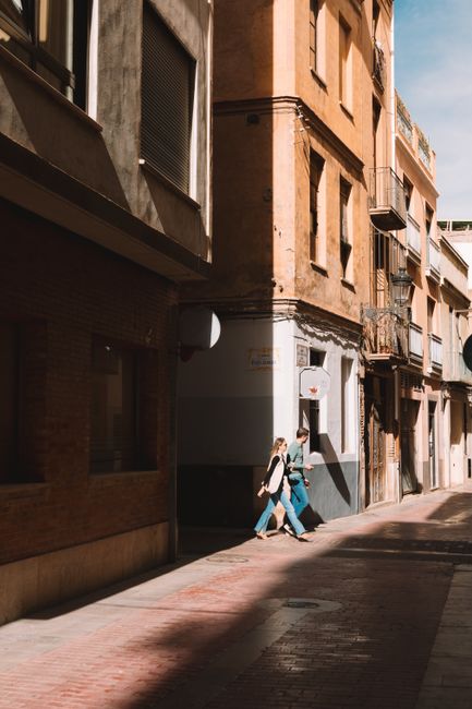 A couple strolls through a sunlit city street with charming old architecture.